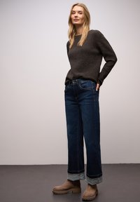 Dunkelgrauer, gerippter Pullover kombiniert mit dunkelblauen Jeans mit weitem Bein und umgeschlagenen Säumen. Das Modell trägt beige Wildleder-Stiefeletten.