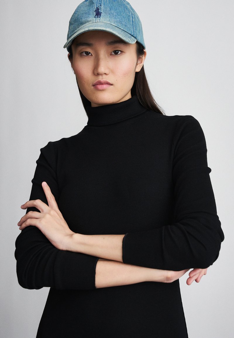 Melna ribota turtlenecka džemperis ar garām piedurknēm, kombinēts ar gaiši zilu džinsa cepuri ar logotipu, izceļot gludu tekstūru.