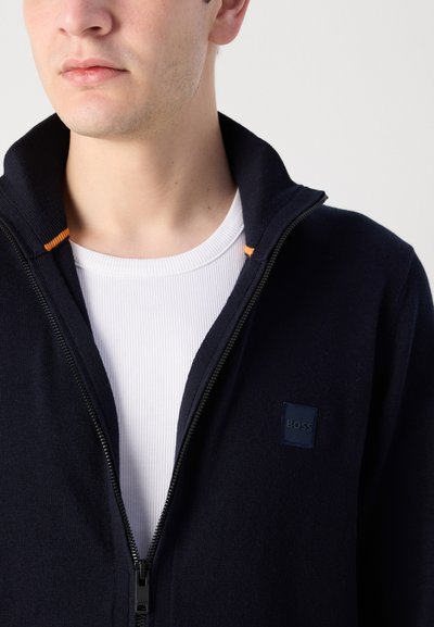 Chaqueta de botones navy con cuello alto, presenta un pequeño parche de logo azul oscuro y un acento naranja en el cuello. Llevada sobre una blusa blanca acanalada.