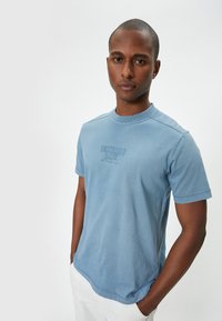 Himmelblaues Baumwoll-T-Shirt mit Rundhalsausschnitt, kurzen Ärmeln und einem dezenten Grafikdruck auf der Vorderseite. Glatte Haptik, lässige Passform.