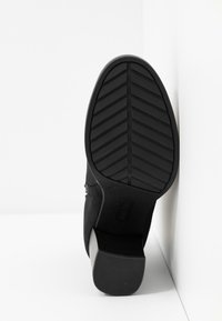 Botte à talon en daim noir avec une semelle en caoutchouc texturée, dotée d'un talon bloc et d'un motif de bande de roulement en zigzag pour une bonne adhérence. Fermeture éclair latérale visible.