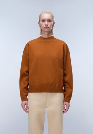 Femme aux cheveux blonds courts portant un pull couleur rouille et un pantalon beige, debout devant un fond gris clair uni.