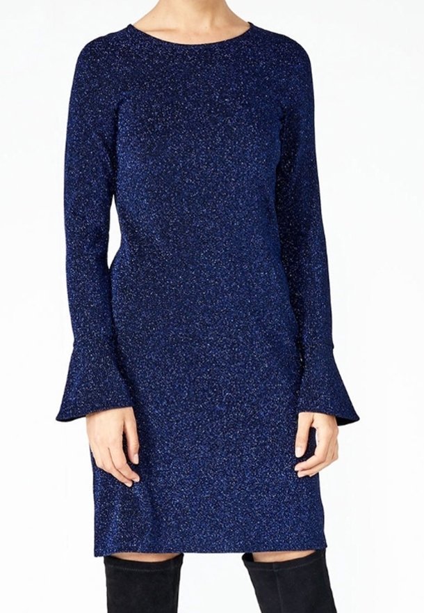 Femme portant une robe longue à manches longues bleu marine scintillante avec des poignets évasés, associée à des bottes noires aux genoux, debout devant un fond uni.