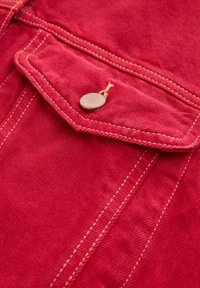 Red cotton fabric with visible stitching, featuring a single pocket and a metal button closure. Texture appears smooth and slightly textured.