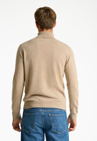 Jersey de punto beige con un cuello alto, puños y dobladillo acanalados. La tela parece suave con una textura sutil, combinado con jeans azules.