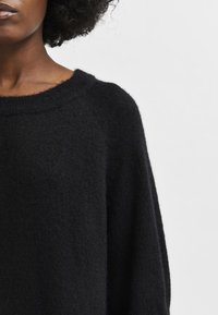 Juodas, minkštas megztinis su plačia iškirpte ir laisvu kirpimu. Turi raglano rankoves ir pūkuotą tekstūrą, suteikiančią jaukų įspūdį.