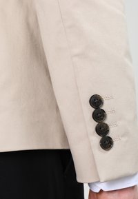 Manga de blazer beige con cuatro botones negros y detalles de costura sutiles. La tela tiene una textura suave y un diseño ajustado.