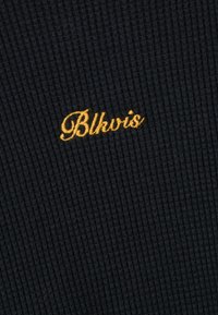 Ribbet svart stoff med synlig tekstur. Har en kursiv gylden brodering av ordet "Blkwis" i nederste høyre hjørne.