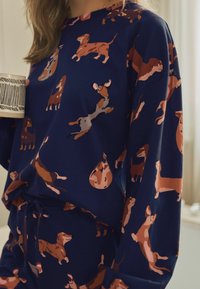 Marineblauwe loungewear met herhaalde teckelpatronen in verschillende bruintinten. Zacht materiaal met een ontspannen pasvorm en een trekkoord in de taille.