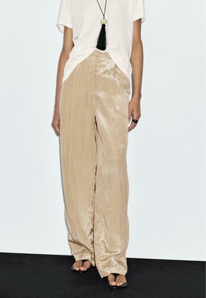 Woman wearing loose, shiny beige wide-leg pants, white short-sleeve shirt, black tassel necklace, black ring, and black open-toe sandals.