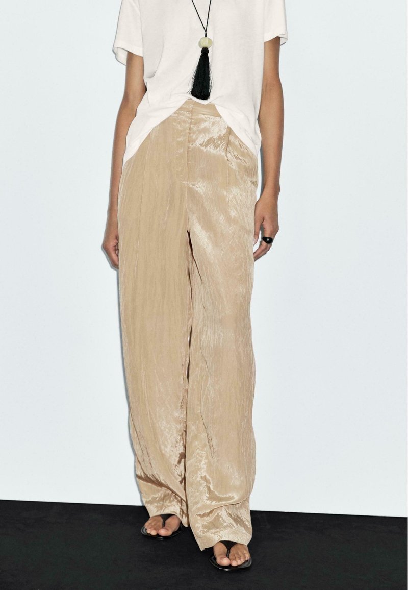 Femme portant un pantalon large beige brillant, une chemise blanche à manches courtes, un collier à franges noir, une bague noire et des sandales noires ouvertes.