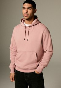 Sudadera con capucha rosa hecha de una tela suave, con un bolsillo tipo canguro en el frente y puños acanalados. Lleva puesta sobre una camiseta blanca con unos jeans negros.
