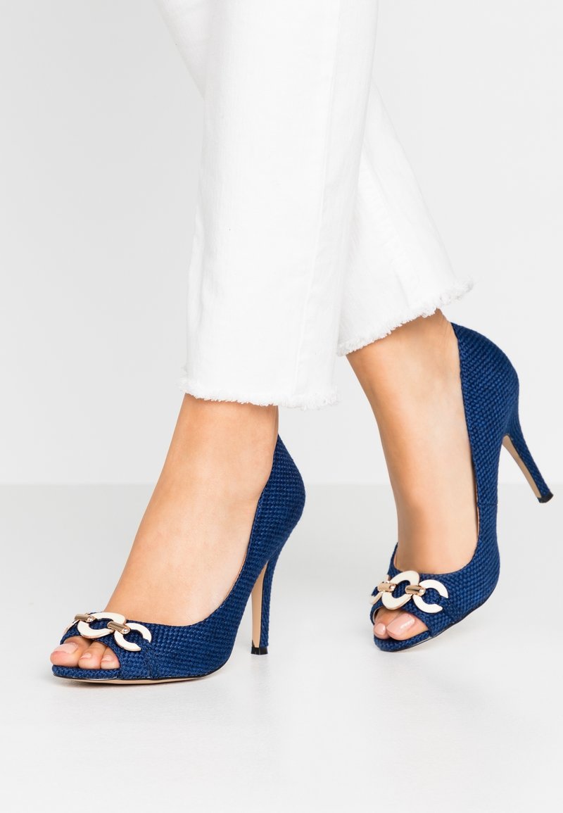 Pieds portant des escarpins à bout ouvert texturés bleus avec des accents de chaînes dorées, associés à un pantalon blanc raccourci à ourlet effiloché.