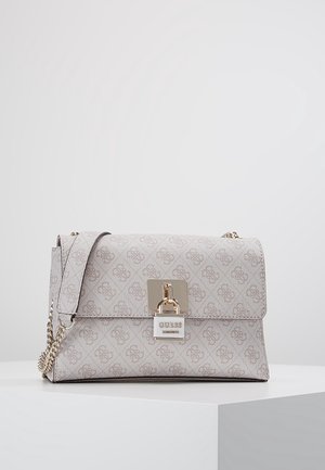 Beżowa torebka na ramię z monogramem, złotym łańcuszkowym paskiem i ozdobnym zamkiem na przedniej klapie, leżąca na białej powierzchni.