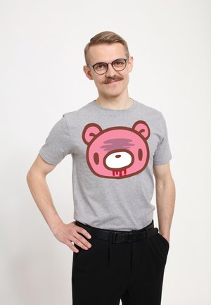 Ung mand med briller og overskæg iført en grå t-shirt med en lyserød bamseansigt-grafik og sorte bukser, stående mod en hvid baggrund.