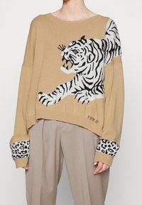 Pull en tricot beige présentant un graphisme de tigre en noir et blanc, des manches larges et des poignets à imprimé léopard. Design léger et court.