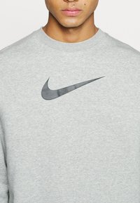 Man som bär en ljusgrå sweatshirt med svart Nike-swoosh-logotyp på bröstet, synlig från axlarna till läpparna.