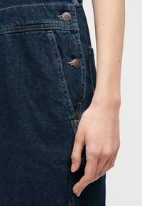 Κοντινό πλάνο ενός χεριού που αναπαύεται δίπλα σε σκουρόχρωμες μπλε ντένιμ σαλοπέτες με ορατά κουμπιά και λεπτομέρειες ραφής.