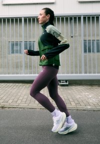 Veste de course verte et noire, leggings violets ajustés, avec des accents réfléchissants et des chaussures de course légères. Cadre urbain pavé.