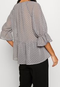 Gennemsigtig grå bluse med hvide prikker, puffærmer og peplumkant. Har en rund halsudskæring og sammenholdte detaljer bagpå.