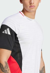 Camiseta deportiva blanca con detalles en negro y rojo, que presenta un patrón texturizado, mangas cortas y un distintivo logo de tres rayas.