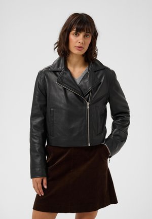 Femme aux cheveux foncés mi-longs, portant une veste en cuir noire par-dessus un haut gris et une jupe marron sur un fond uni.