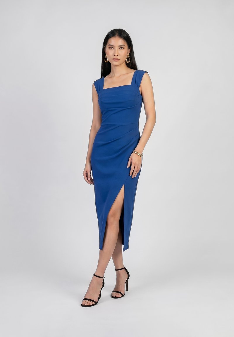 Femme en robe midi bleue sans manches avec une fente à l'avant, portant des sandales noires à talons hauts, des boucles d'oreilles créoles dorées et un bracelet, debout.