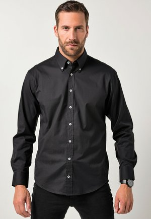 Mann mit kurzen Haaren und Bart, der ein schwarzes langärmliges Hemd mit Knöpfen und schwarze Hose vor einem schlichten hellen Hintergrund trägt.