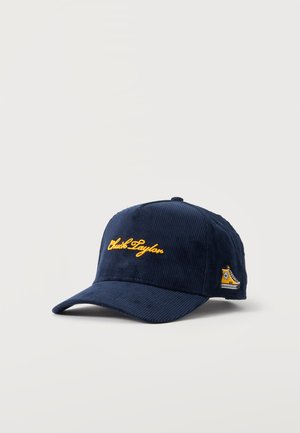 Cappellino da baseball in velluto a coste blu scuro con testo giallo corsivo sul davanti e piccola toppa gialla a forma di sneaker sul lato.