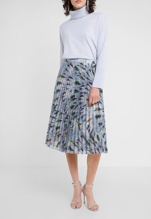 Femme portant un col roulé bleu clair, une jupe midi plissée à motif abstrait, des sandales argentées à talons hauts et de longues boucles d'oreilles pendantes.