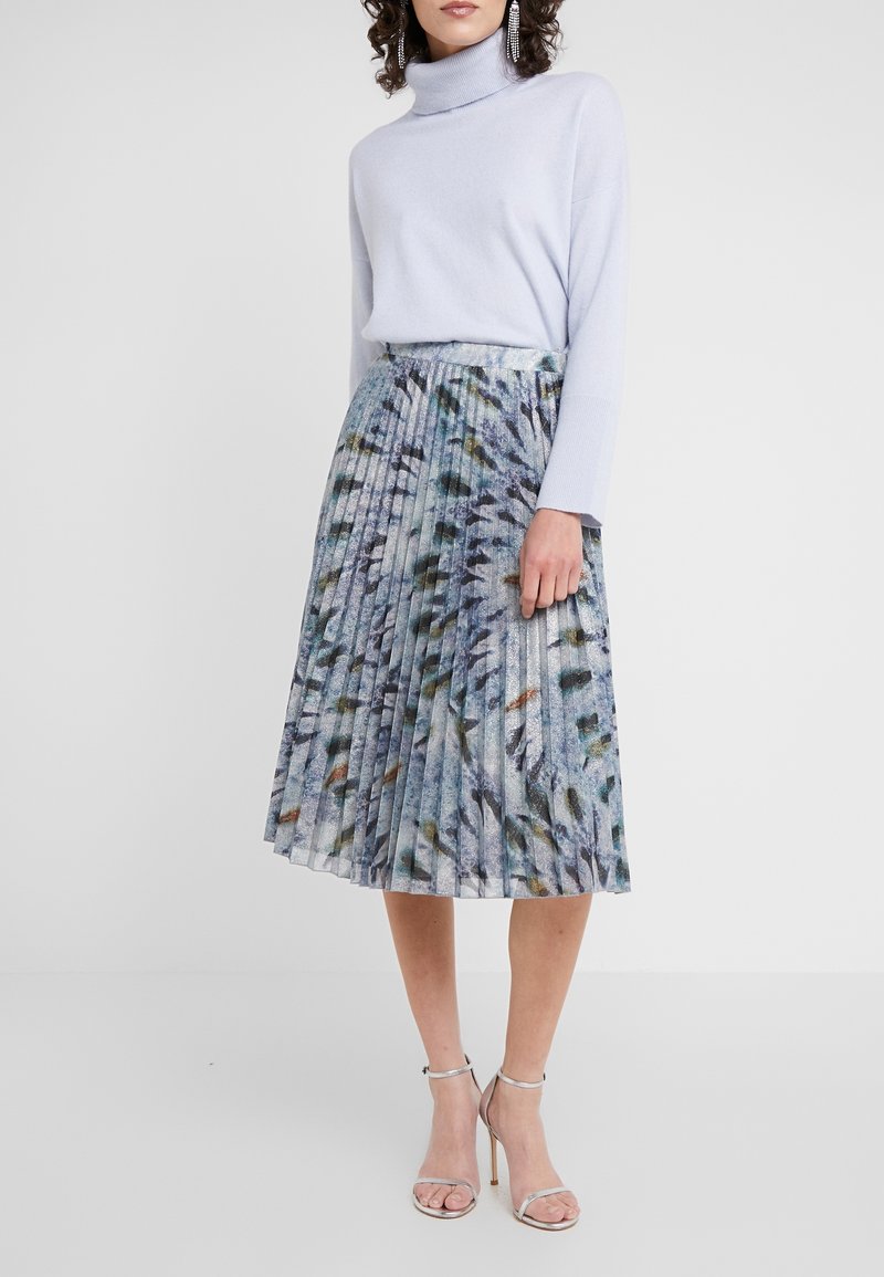 Femme portant un col roulé bleu clair, une jupe midi plissée à motif abstrait, des sandales argentées à talons hauts et de longues boucles d'oreilles pendantes.