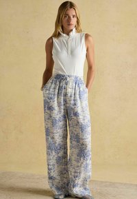 Bezramenná bílá blůza s vysokým límcem; volné, vzorované modro-bílé kalhoty s širokými nohavicemi, elastickým pasem a kapsami.