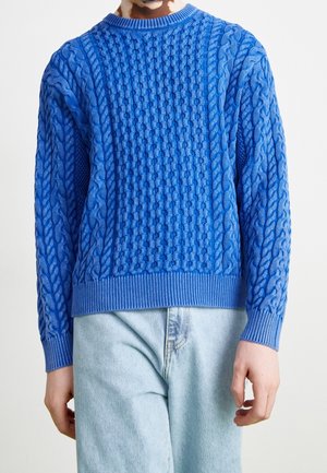 Henkilö, joka pukeutunut kirkkaan siniseen palmikkoneulepuseroon, jossa on pitkät hihat, ja vaaleansiniset farkut, kuvattuna kaulasta lantiolle yksiväristä taustaa vasten.