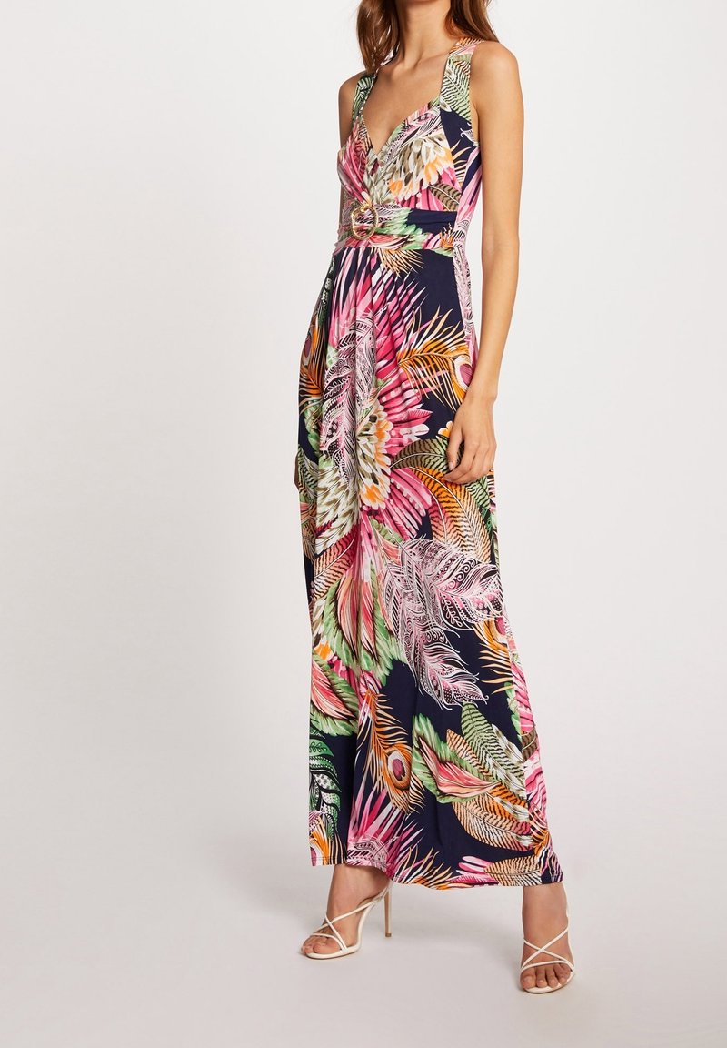 Femme portant une robe longue sans manches à col en V avec ceinture, imprimé plumes tropicales rose, orange, vert et bleu marine, et des sandales blanches à talons hauts à brides.