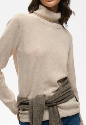 Kvinde iført en beige rullekravesweater med en mørkegrå sweater bundet om taljen, kombineret med blå jeans.
