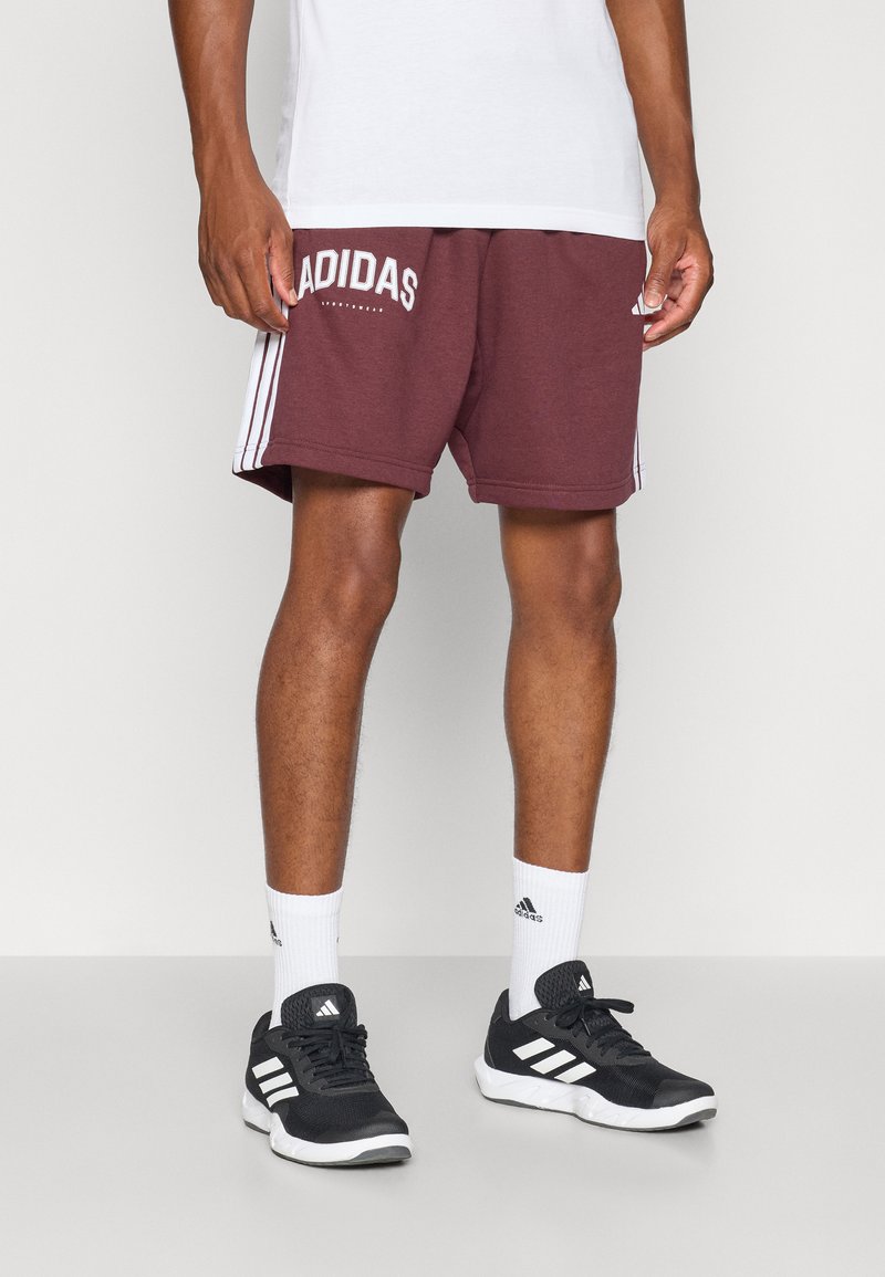 Pantalones cortos deportivos color burdeos con el logo blanco de Adidas y tres rayas en los lados, combinados con zapatillas negras y calcetas blancas.
