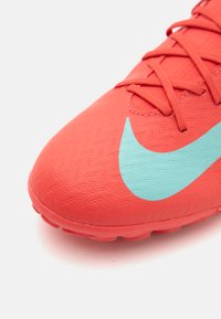 Nike fodboldsko i lys koralfarve med en struktureret overflade. Har en turkise swoosh og koordinerede snørebånd, designet til ydeevne.
