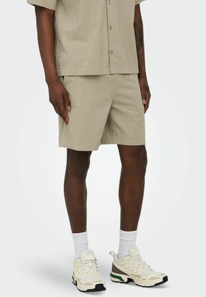 Mann iført beige teksturerte shorts og matchende kortermet skjorte, hvite sokker og beige treningssko med grønne og svarte detaljer.