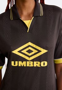 Čierna polo tričko so žltými akcentmi, obsahujúce textúrovaný geometrický vzor a výrazné žlté logo "UMBRO" vpredu.