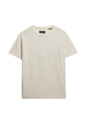 T-shirt corto a maniche corte color beige chiaro, realizzato in morbido cotone, con un design di testo bianco in rilievo disposto in modo circolare, con collo tondo.