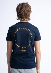 Marineblauwe katoenen t-shirt met korte mouwen, met een cirkelvormige oranje grafische tekst op de achterkant, met de tekst "PETROL INDUSTRIES" en projectdetails eronder.