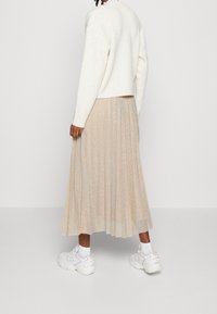 Osoba w białym prążkowanym swetrze, beżowej plisowanej spódnicy midi i białych sneakersach, stojąca na jednolitym jasnym tle.