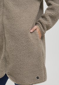 Beige fleece jack met een zachte, gestructureerde oppervlakte, voorzijde rits, zijzakken en contrasterende biezen langs de randen.