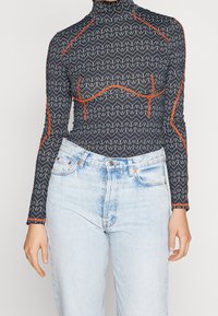 Långärmad svart topp med vitt zigzag-mönster, hög krage och orange detaljer i sömmen, matchad med ljusblå jeans med hög midja.