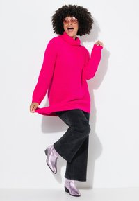Dicker Strickpullover in leuchtendem Pink mit einem Rollkragen, kombiniert mit schwarzen, verwaschenen Schlagjeans und glänzenden, lavendelfarbenen Ankle-Boots.