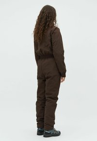 Salopetă maro, impermeabilă, cu talie elastică, mâneci lungi și manșete strânse, asortată cu încălțări robuste pentru drumeții, văzută din spate.