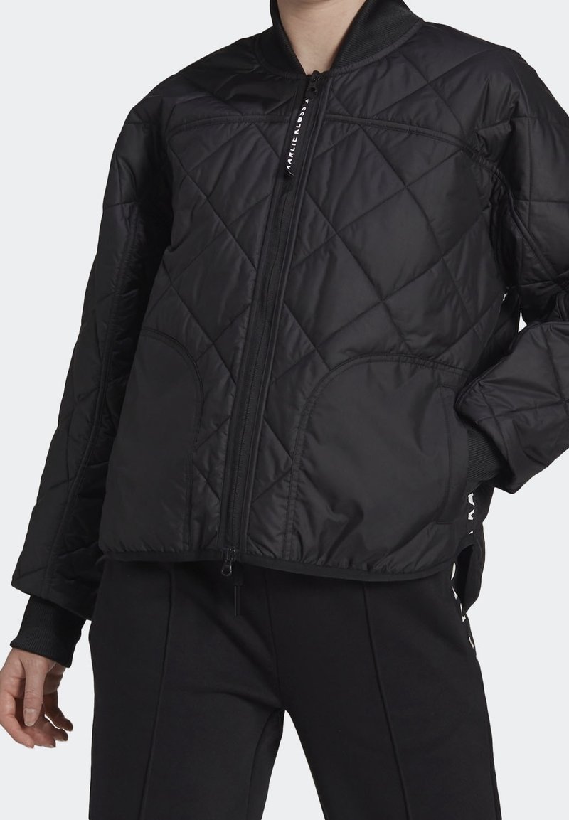 Personne portant une veste matelassée noire avec une fermeture éclair frontale et des poignets côtelés, associée à un pantalon noir sur un fond uni.