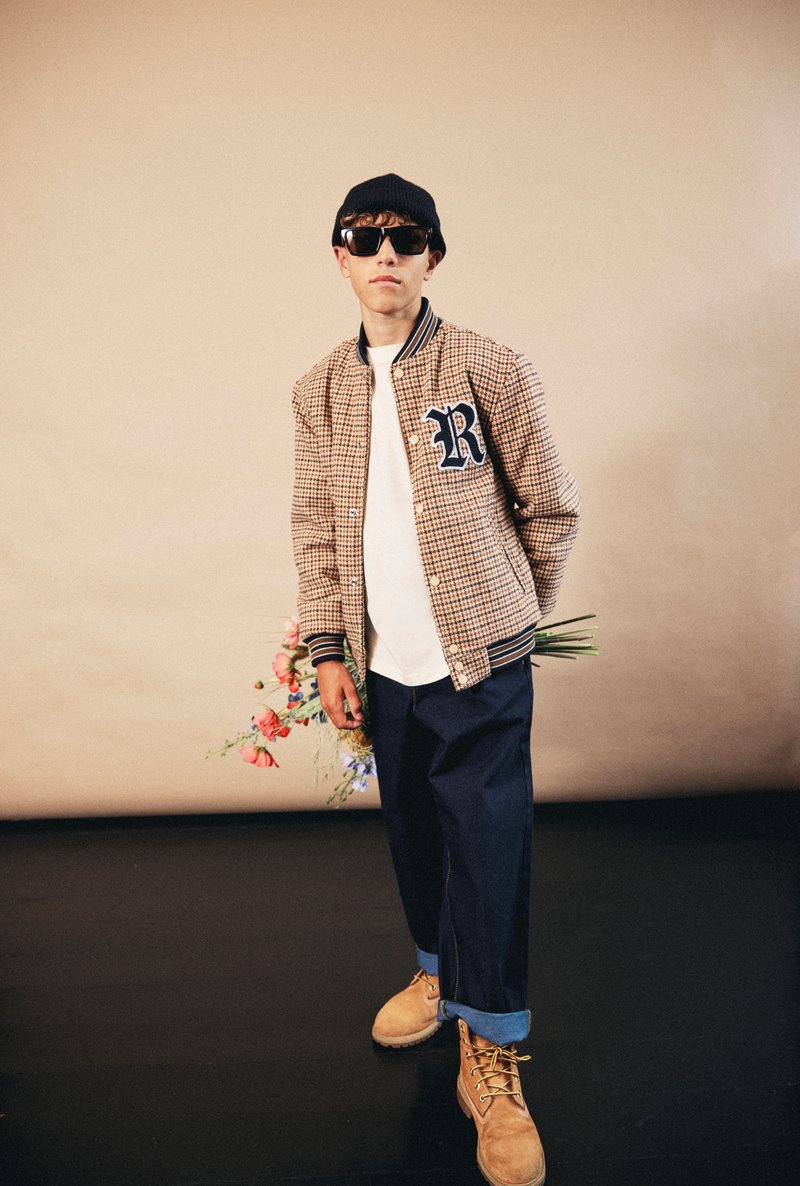 Veste bomber en houndstooth brun avec des accents bleu marine, t-shirt blanc, pantalons larges bleu foncé et bottes beiges, tenant des fleurs.