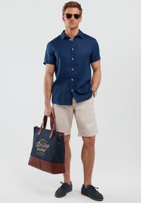 Camicia blu di lino a maniche corte, pantaloni corti beige, borsa a tracolla blu navy con dettagli in pelle marrone, occhiali da sole e scarpe blu navy. Design semplice e vestibilità casual.