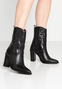 Bottines en cuir noir avec des talons blocs hauts, bouts pointus, fermeture éclair latérale et des coutures décoratives portées sur des jambes nues.